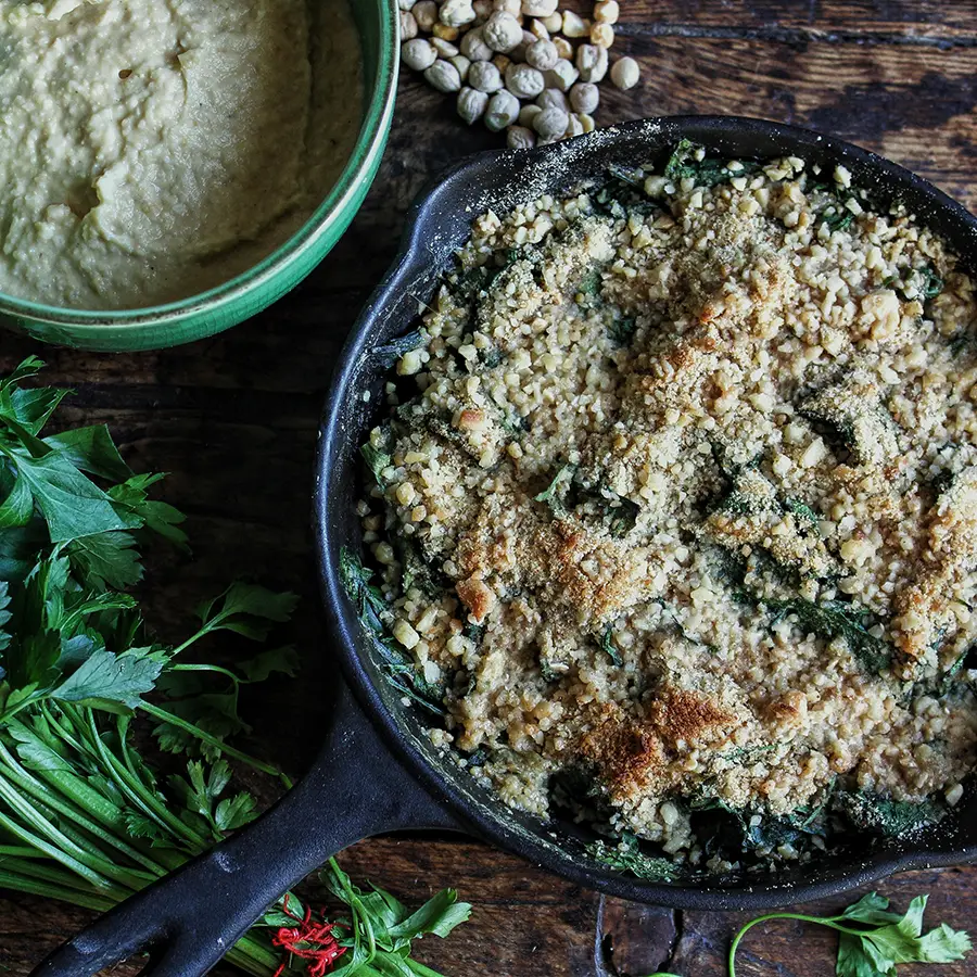 A frying pan with food and a bowl of hummus on a dark brown table with herbs around it.